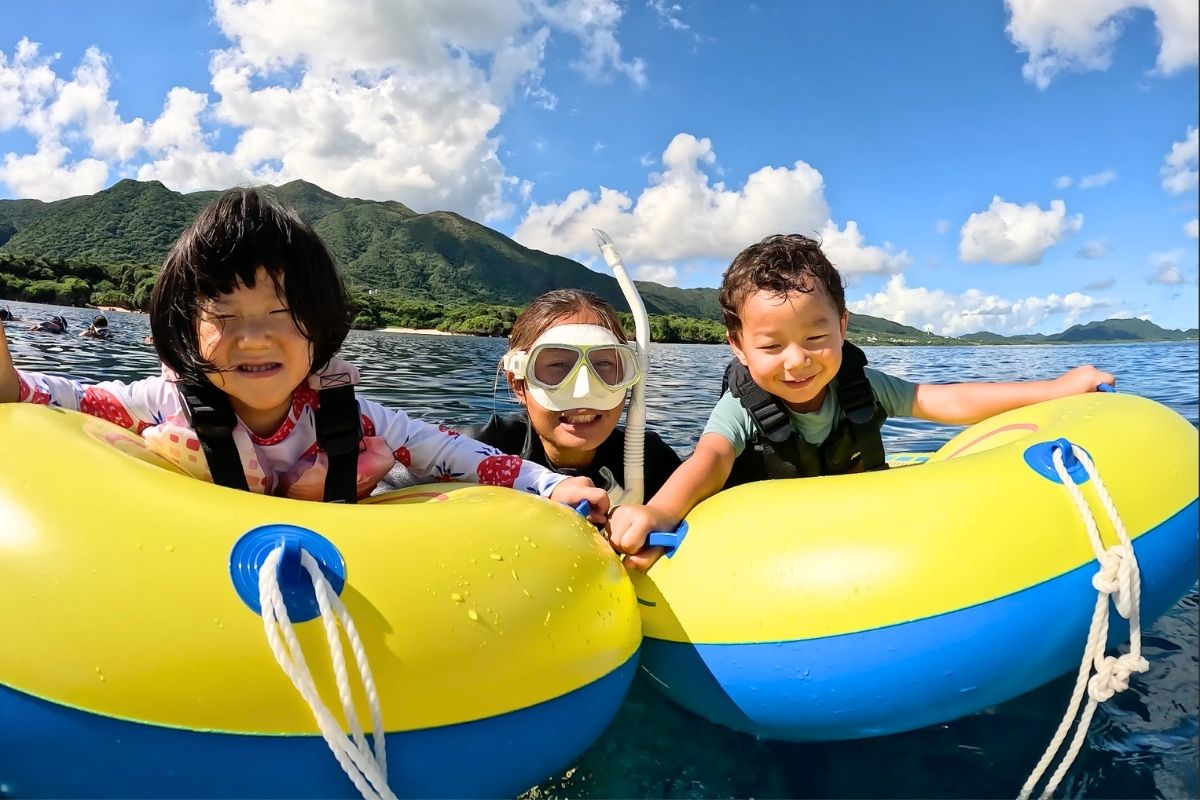 石垣島のきれいな海で黄色い浮き輪に乗って満面の笑顔を見せる小さな子供たちとガイドスタッフ