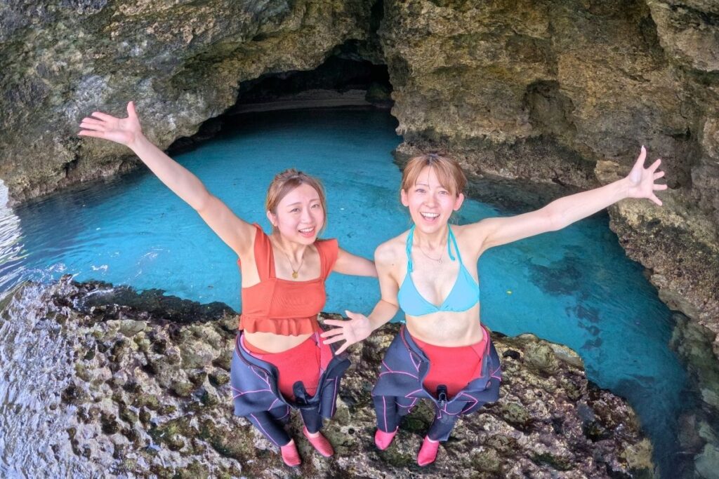 神秘的なエメラルドグリーンの水面が広がる石垣島・青の洞窟の前で両手を広げて満面の笑顔を見せる水着姿の女性2人組