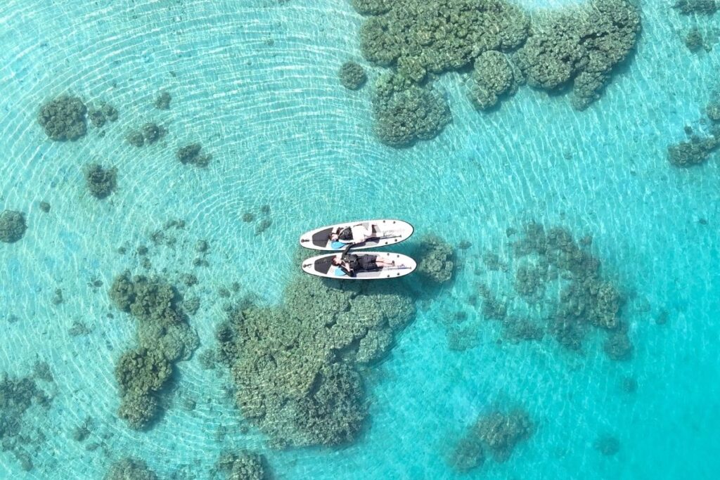 底まで透き通った石垣島の浅瀬で、並べたサップボードの上に大の字になって寝転びリラックスする様子を真上から捉えたドローン写真