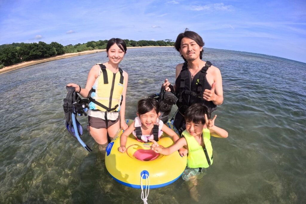 浅瀬のきれいな海で黄色い浮き輪チューブを囲んで楽しむ子供連れご家族のシュノーケリング体験風景