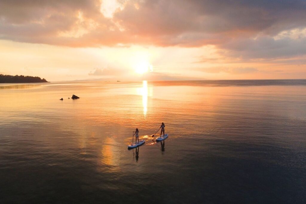 どこまでも続く黄金色の海をドローンで広角空撮した、夕日へ向かってパドルを漕ぐ2人のサンセットSUP絶景フォト