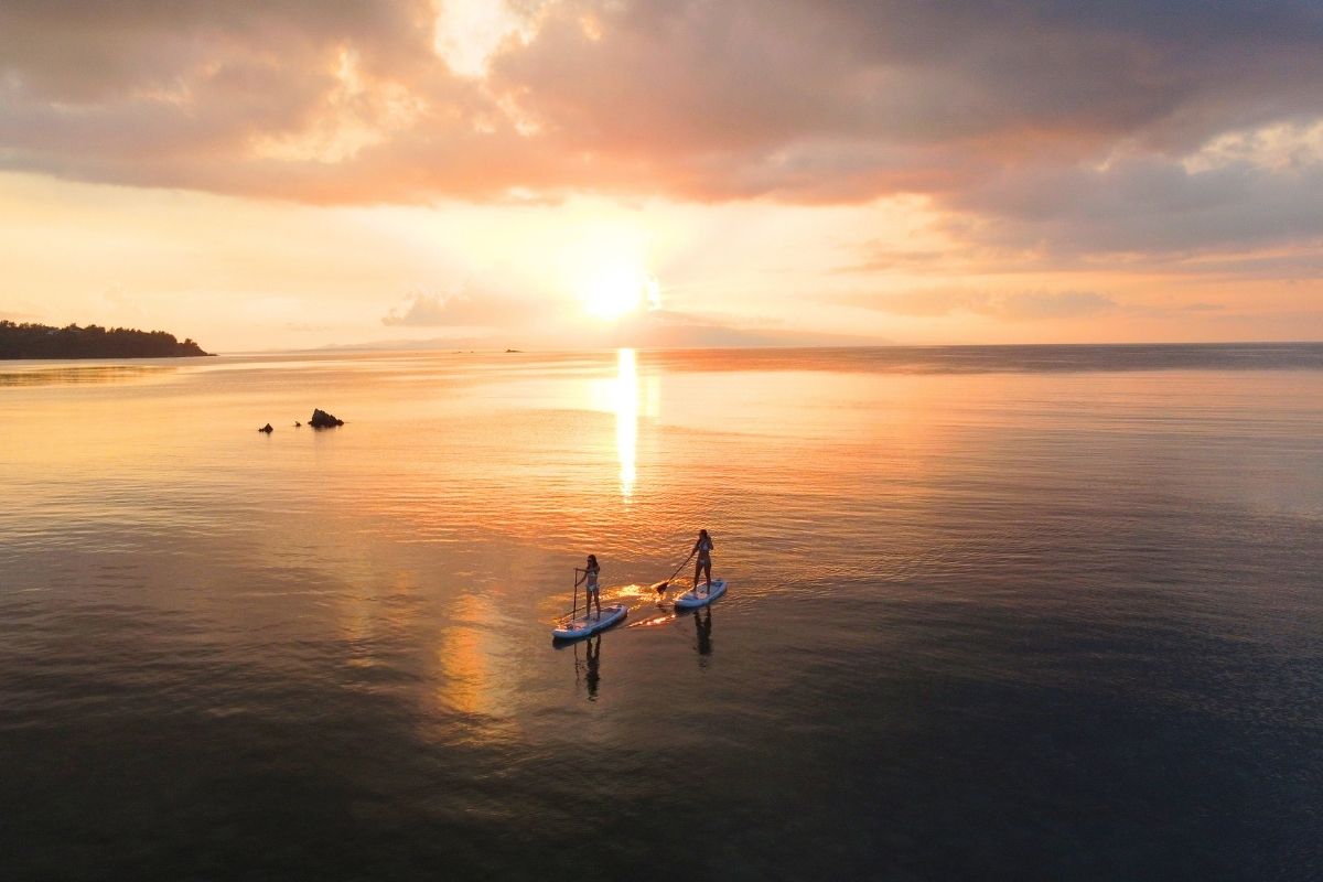 どこまでも続く黄金色の海をドローンで広角空撮した、夕日へ向かってパドルを漕ぐ2人のサンセットSUP絶景フォト