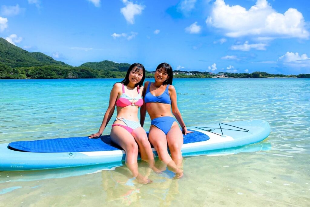 リゾート感あふれる石垣島の澄み切った浅瀬で、サップボードの上に仲良く腰掛けて笑顔を見せる水着姿の女性2人組